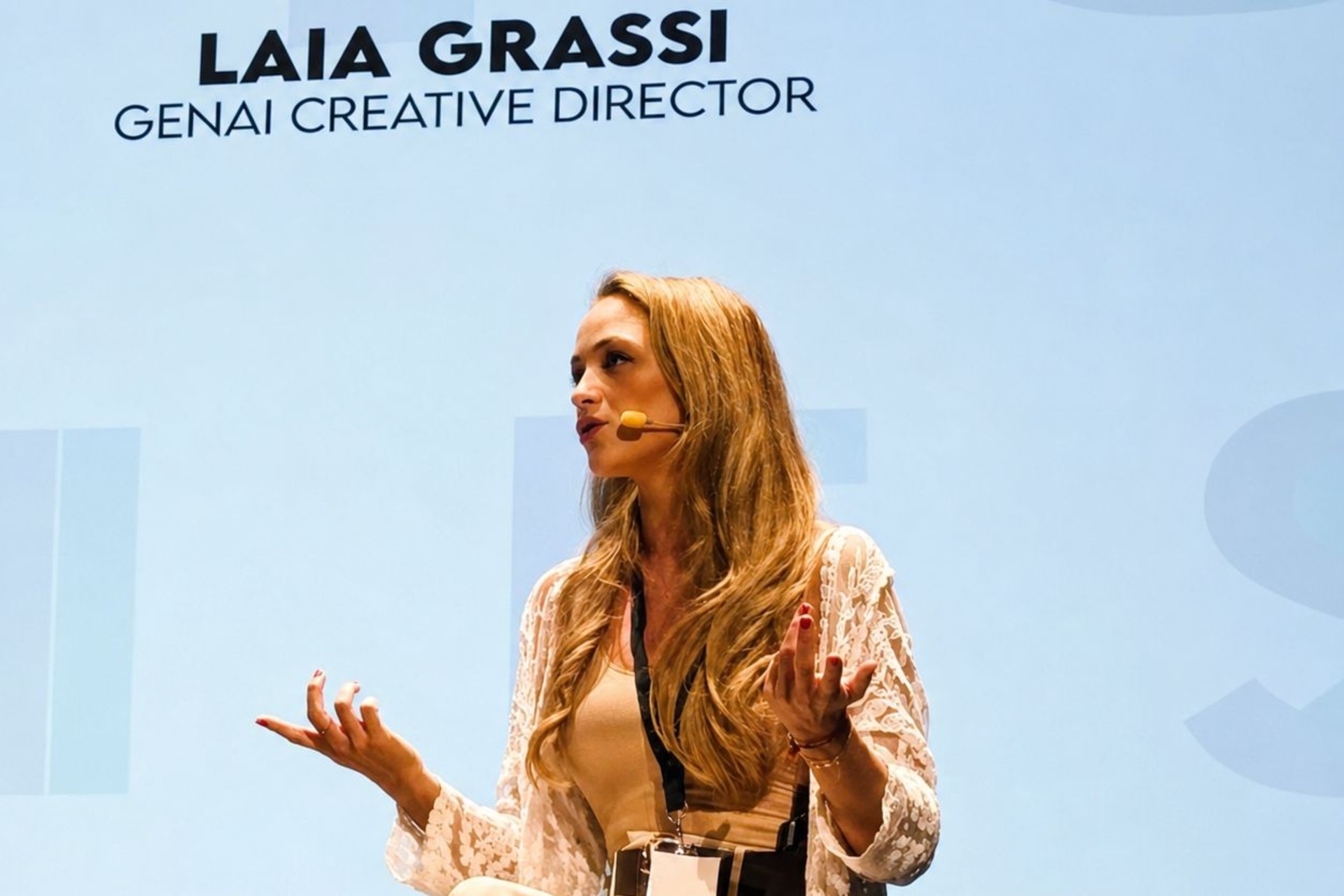 Laia Grassi: pembicara di atas panggung selama acara yang didedikasikan untuk kecerdasan buatan generatif, kreativitas, inovasi, dan arahan kreatif, dengan layar di belakangnya dan mikrofon headset yang dikenakan.